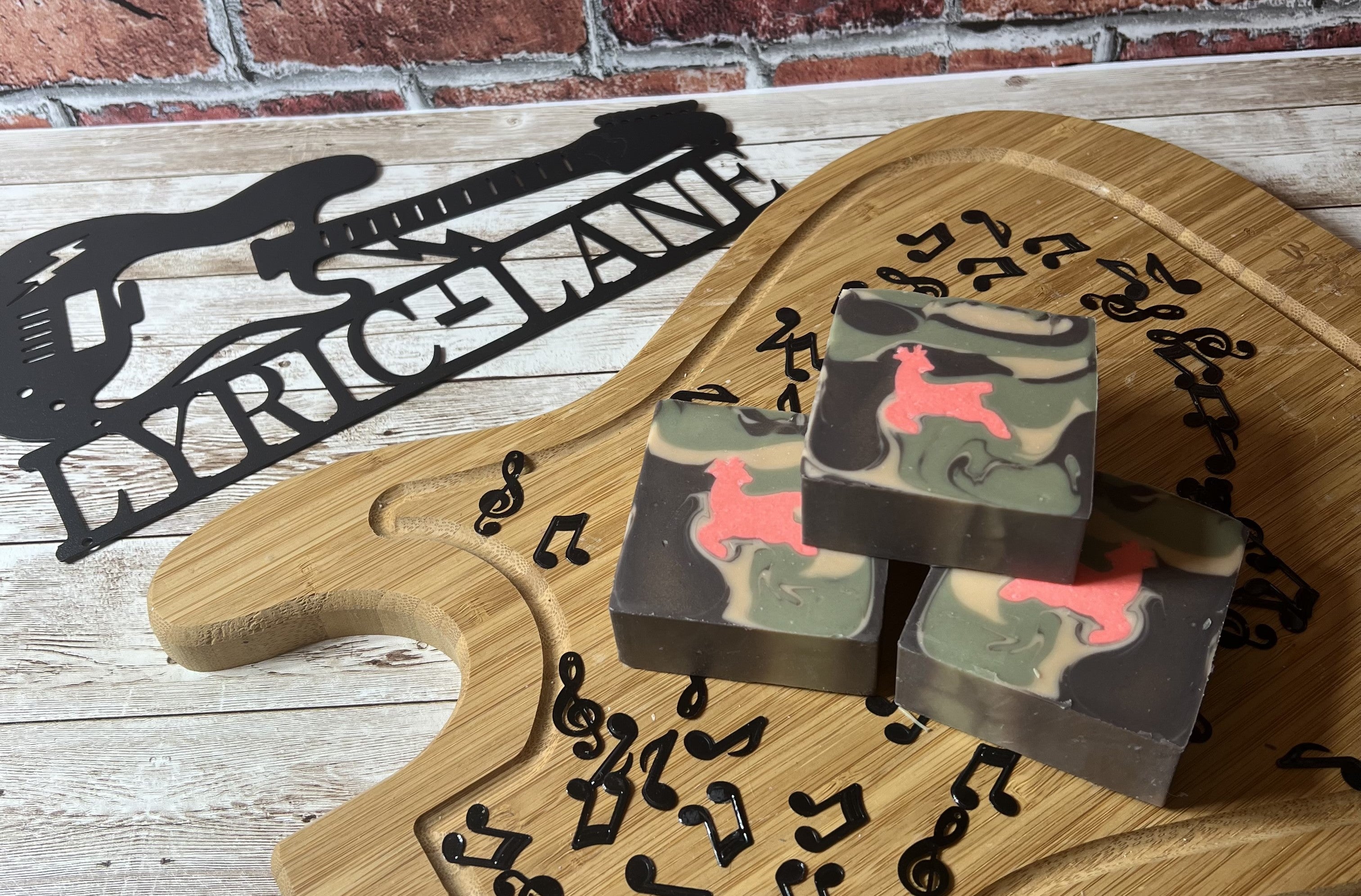 Three marbled soap bars with pink animal shapes on guitar-shaped wooden board, featuring Another 1 Bites the Dust - Smells Like Dirt. On Purpose.