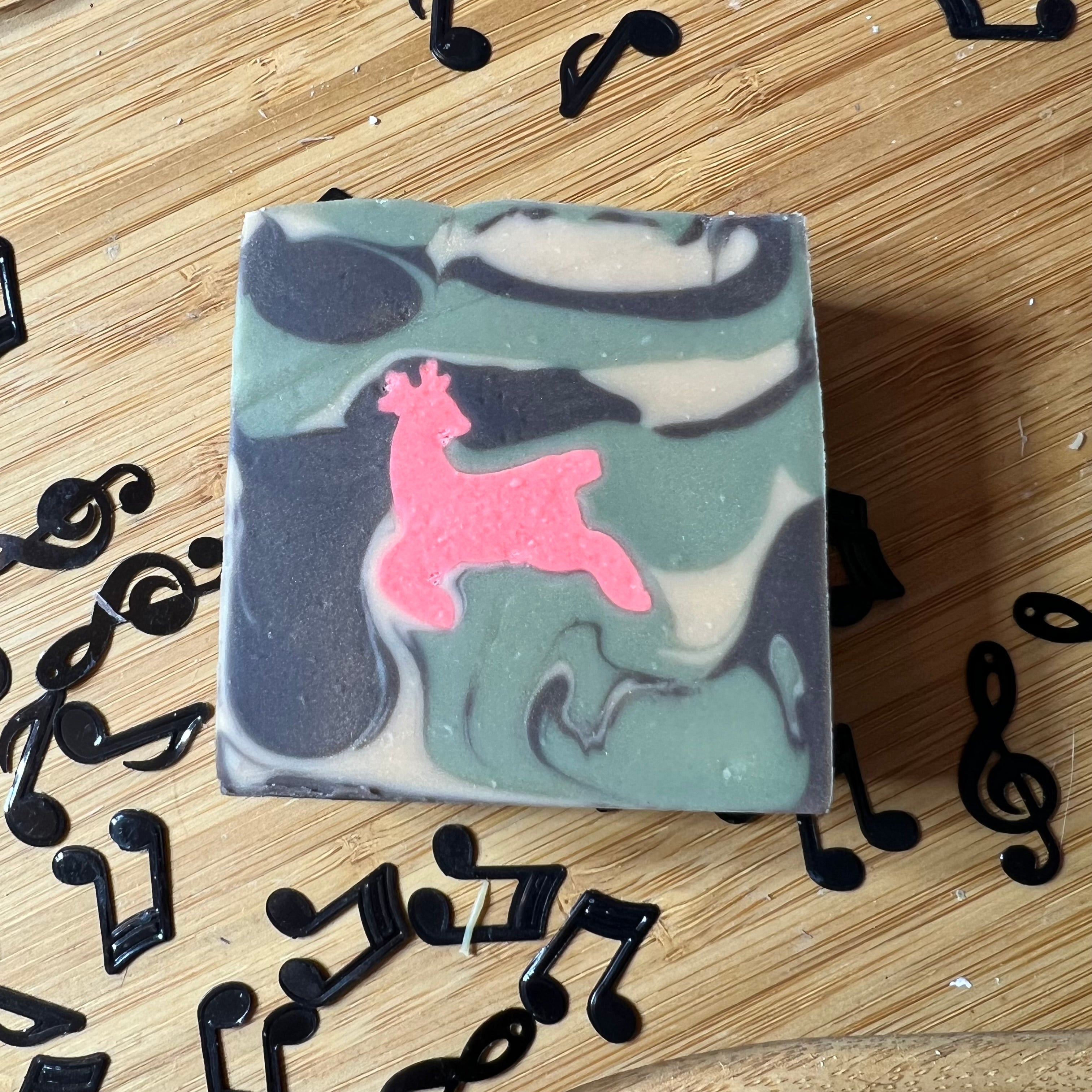 Square soap bar with green and gray swirls and a pink animal shape, surrounded by black music note confetti, featuring Another 1 Bites the Dust - Smells Like Dirt. On Purpose. by Lyric-Lane