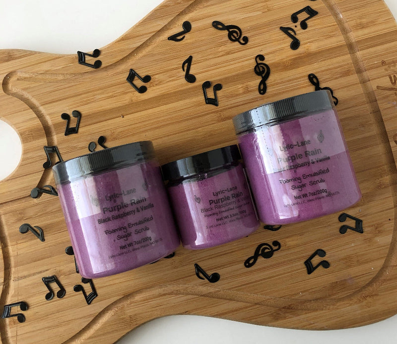 Three jars of Purple Rain-Black Raspberry & Vanilla scented whipped emulsified sugar scrub. Two 7 oz PET plastic jars and one 3.5 oz jar in the middle on a wood guitar shape with scattered music notes. Made by Lyric-Lane.