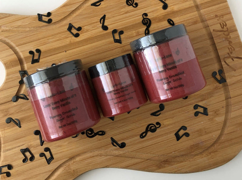 Three jars of crimson Lovin' Every Minute of It-Berry Vanilla scented whipped emulsified sugar scrub. Two 7 oz PET plastic jars and one 3.5 oz jar in the middle on a wood guitar shape with scattered music notes. Made by Lyric-Lane.