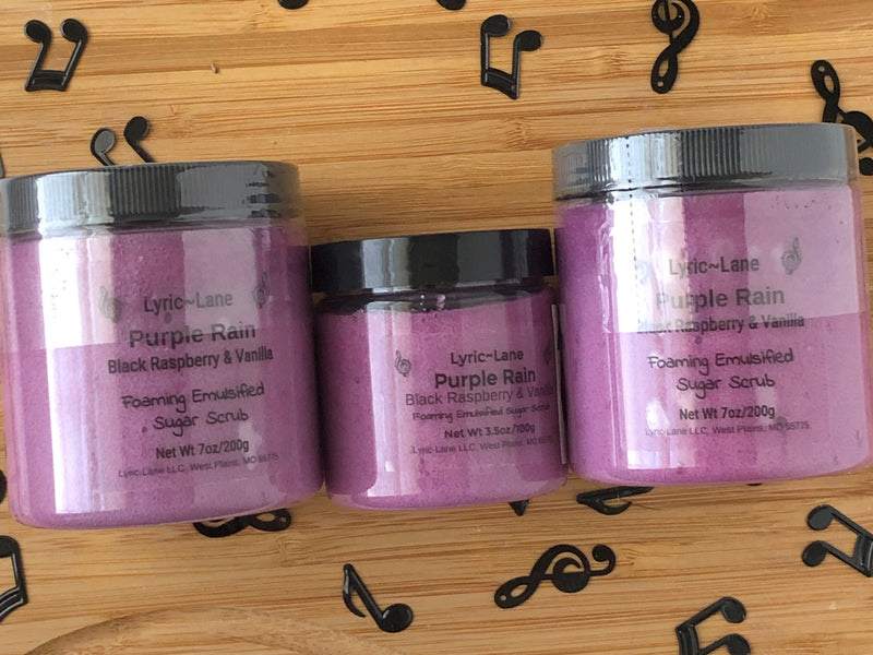 Three jars of Purple Rain-Black Raspberry & Vanilla scented whipped emulsified sugar scrub. Two 7 oz PET plastic jars and one 3.5 oz jar in the middle on a wood background with scattered music notes. Made by Lyric-Lane.