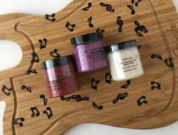 Three varieties of whipped emulsified sugar scrubs scented with crimson Berry Vanilla, purple Black Raspberry Vanilla, and white Fresh Vanilla in 3.5 oz, 100g PET plastic jars on a wood guitar shape with scattered music notes. Made by Lyric-Lane