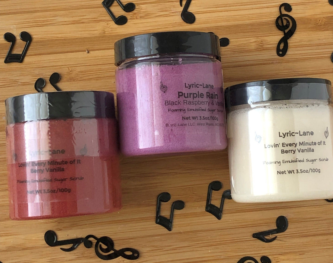 Three varieties of whipped emulsified sugar scrubs scented with crimson Berry Vanilla, purple Black Raspberry Vanilla, and white Fresh Vanilla in 3.5 oz, 100g PET plastic jars on a wood guitar shape with scattered music notes. Made by Lyric-Lane