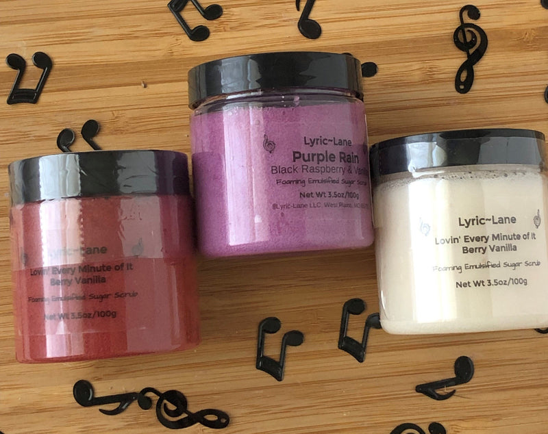 Three varieties of whipped emulsified sugar scrubs scented with crimson Berry Vanilla, purple Black Raspberry Vanilla, and white Fresh Vanilla in 3.5 oz, 100g PET plastic jars on a wood guitar shape with scattered music notes. Made by Lyric-Lane