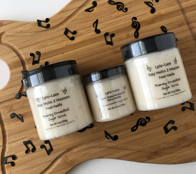 Three jars of white Say Hello 2 Heaven-Fresh Vanilla scented whipped emulsified sugar scrub. Two 7 oz PET plastic jars and one 3.5 oz jar in the middle on a wood guitar shape with scattered music notes. Made by Lyric-Lane.