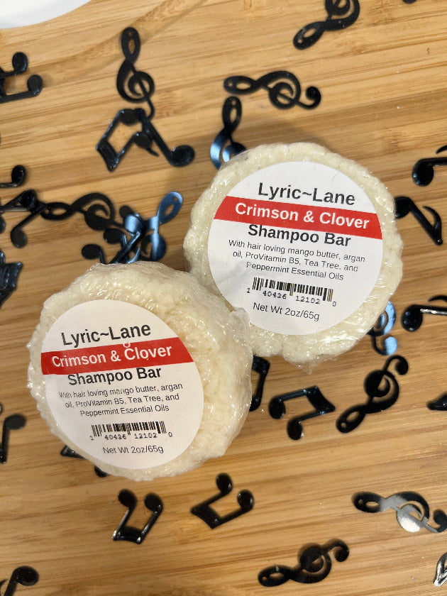 Two 2oz round bars of Crimson and Clover Shampoo Bars. White bars scented with tea tree and peppermint essential oils on a wood guitar shape with scattered music notes. Made by Lyric-Lane