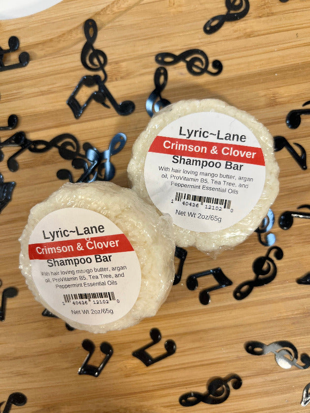 Two 2oz round bars of Crimson and Clover Shampoo Bars. White bars scented with tea tree and peppermint essential oils on a wood guitar shape with scattered music notes. Made by Lyric-Lane