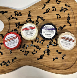 Four different 2oz round shampoo bars arranged in an arc on a wood guitar shape with scattered music notes. One each of Hotel California, Crimson and Clover, Beautiful Believer, and Say Hello 2 Heaven. Made by Lyric-Lane