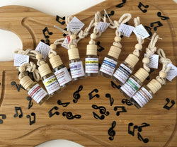Nine different scents in 7.5 ml glass diffuser bottles with an absorbent wooden top and attached ropes for hanging displayed on a wood guitar shape with scattered music notes. Made by Lyric Lane Soap.